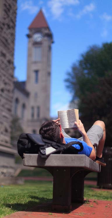 A student reads a book while lying on a bench.
